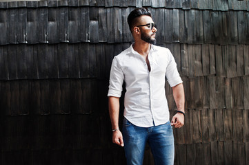 Obraz premium Stylish tall arabian man model in white shirt, jeans and sunglasses posed against wooden wall indoor. Beard attractive arab guy.