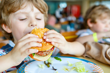 Cute healthy preschool kid boy eats hamburger sitting in school or nursery cafe. Happy child eating...