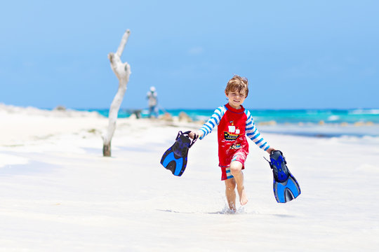 Adorable little blond kid boy having fun on tropical beach of Maldives. Excited child playing and surfing in sun protected swimsuit in ocean on vacations. White sand, Kid holding flippers for swimming