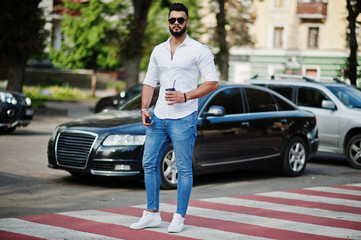 Stylish tall arabian man model in white shirt, jeans and sunglasses posed at street of city. Beard attractive arab guy with cup of coffee against black business car.