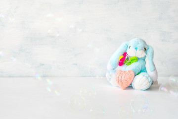 Cute toy soft blue bunny with a flower and pink thread heart with soap bubbles on light background. Birthday card, Valentine's Day, mother's day, Easter background.