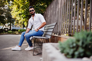 Fototapeta premium Stylish tall arabian man model in white shirt, jeans and sunglasses posed at street of city. Beard attractive arab guy sitting on bench.