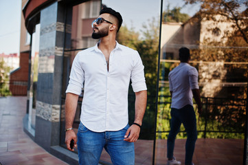 Stylish tall arabian man model in white shirt, jeans and sunglasses posed at street of city. Beard attractive arab guy against modern building.