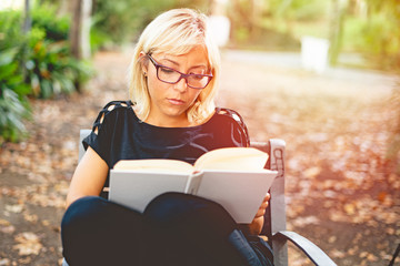 Young adult blonde girl reading a book outdoor in a park chair. Blank white book cover