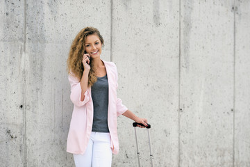 Woman using smart phone and holding luggage while standing against the wall.