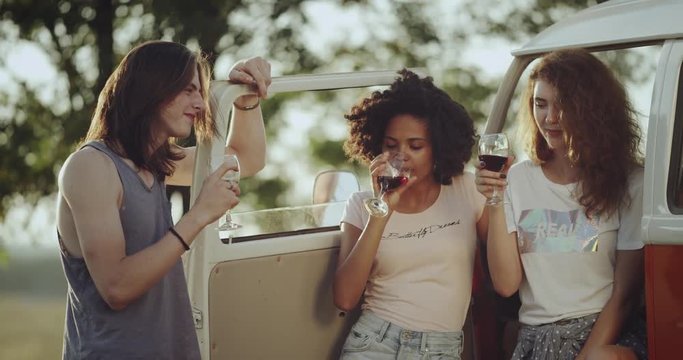 Three friends have a picnic time , drinking wine and smiling have a good mood in the middle of nature standing beside of a retro bus.