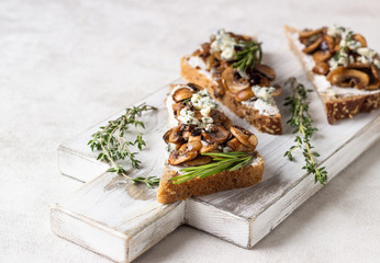 Whole grain rye bread toast with cream cheese or ricotta, mushroom and herbs on a white wooden cutting board.