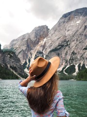 A girl with long hair and a shirt looks directly in front of her at the azure mountain lake in the Dolomites. The girl holds her hand brown wide-brimmed hat