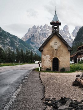 A picturesque panorama in the Dolomites. There are huge mountains covered with snow, coniferous forests, mountain clear azure lake, a lonely wild place, a deserted Church on the road