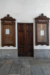 Antica porta con due antiche bacheche in vetro e legno per esposizione pubblicazioni di futuri matrimoni appese su una parete interna bianca in una chiesa - 221427508