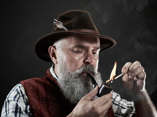 dramatic portrait of gray bearded senior man in hat smoking tobacco pipe. view of Austrian, Tyrolean, Bavarian old man in national traditional costume in retro style.