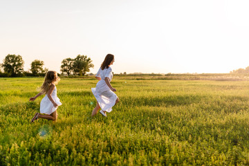mother and daughter running in green meadow with sunset on background