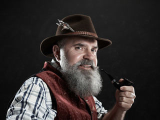 dramatic portrait of gray bearded senior man in hat smoking tobacco pipe. view of Austrian, Tyrolean, Bavarian old man in national traditional costume in retro style.