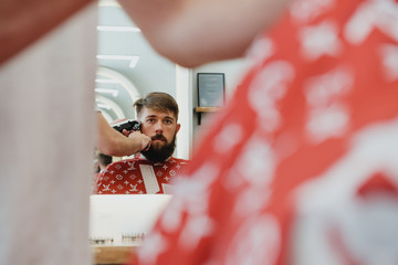 Handsome bearded man in barbershop. Barber cuts hair with  electric trimmer.