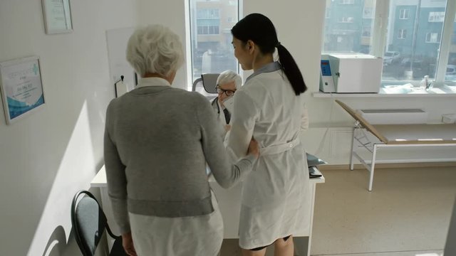 Senior practitioner examining x-ray images while female nurse walking with elderly woman for medical checkup