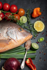 Fresh raw red snapper fish with spices, herbs and vegetables for cooking on dark background. Top view. Vertical composition.