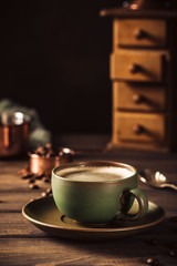 Green cup of coffee with beans and coffee mill on old wooden background with copy space. Retro style toned dark photography.