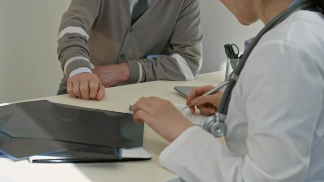 Elderly man sitting at desk in doctor’s office and talking to female specialist while she working with x-ray image and writing down prescription