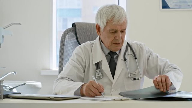 Tracking shot of senior male doctor sitting at desk in the office at clinic, writing down prescription and reading x-ray image