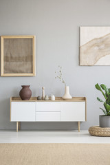 Fototapeta premium White wooden cupboard with decor and vase with flowers standing in real photo of bright room interior with grey wall and posters with fabrics