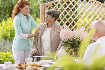 A tender caretaker assisting an elderly woman with a walker during an afternoon snack time on a patio in the garden of a private nursing home.