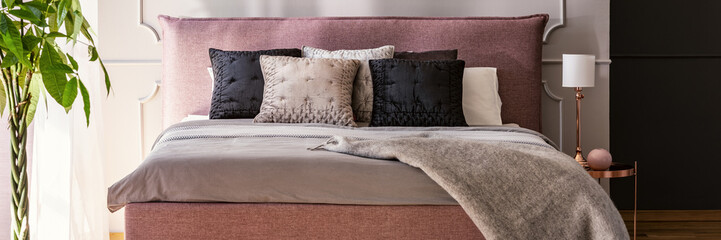 Dirty pink king-size bed with cushions in real photo of grey bedroom interior with rose gold lamp and bedside table