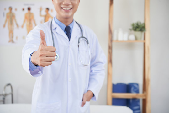 Anonymous man in medical apparel smiling and showing thumb-up gesture to camera while standing in office
