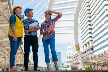 Three industrial engineer wear safety helmet and holding tablet engineering working and talking with drawings inspection on building outside. Engineering tools and construction