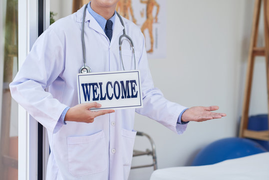 Unrecognizable man in medical apparel holding sign with welcome writing and gesturing with hand while standing in office