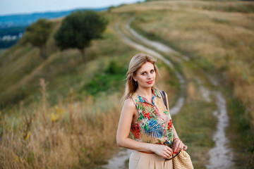 Young beautiful thoughtful woman in tropical blouse with wicked bag standing on village road and looking at camera with smile. Outdoor shot, nature shot, sunny day.