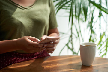 Anonymous female sitting at cafe table near mug of hot drink and browsing modern smartphone