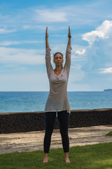 Fototapeta na wymiar Beautiful young girl in black leggings and tunic makes yoga practice on a beach of the ocean with green grass, yellow sand, azure water, blue sky, clouds. Wellness healthy sport photo of female model
