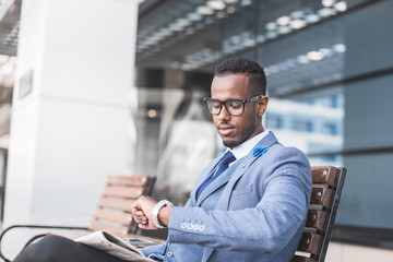 black man businessman in a business suit and glasses sits on a bench and reads a newspaper, looks at the clock against the backdrop of a modern city