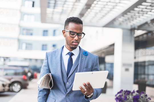 black man businessman in a business suit, expensive watch and glasses with a newspaper and digital tablet goes against the backdrop of a modern city to work
