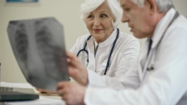 Tilt up of beautiful senior female doctor sitting at desk in clinic and discussing x-ray image with elderly male colleague