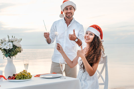 happy couple in santa hats showing thumbs up and celebrating christmas with sparklers on beach