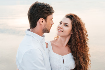 smiling romantic couple looking at each other and hugging on seashore