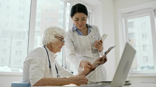 Professional female doctor in lab coat holding digital tablet and explaining x-ray image to senior female colleague while working in medical clinic