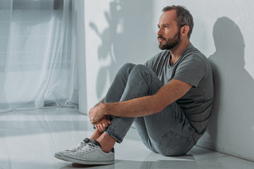 side view of sad bearded mid adult man sitting on floor and looking away