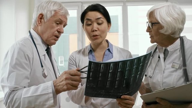 Tilt up of Asian female doctor looking at x-ray image and discussing diagnosis with senior colleagues in clinic