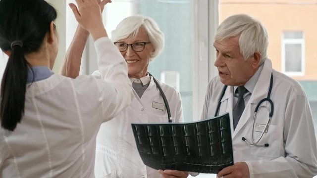 Cheerful senior doctors holding x-ray image, talking to female intern, smiling and giving high five