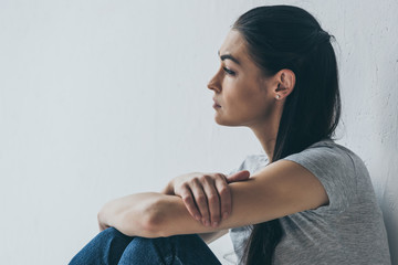side view of beautiful frustrated brunette girl sitting and looking away on grey
