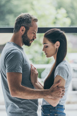 side view of bearded man embracing sad young girlfriend while standing together near window