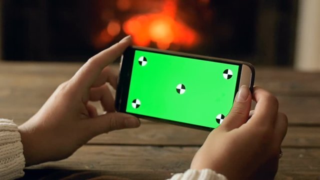 Closeup slow motion footage of female hand holding smartphone against burning fireplace. Green chromakey display for inserting your image or video on screen