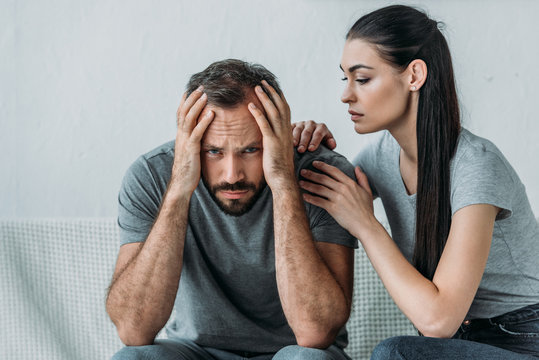 Young Woman Supporting Depressed Frustrated Boyfriend Sitting On Couch And Looking At Camera