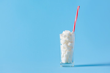 Glass full of sugar cubes with straw isolated on blue background. Unhealthy diet concept. Copy space
