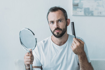middle aged man holding mirror with comb and looking at camera, hair loss concept