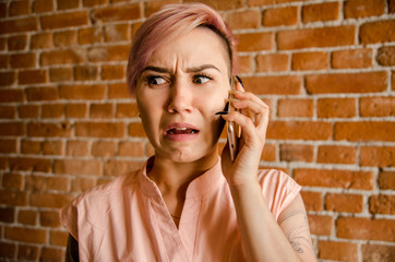 Young beautiful frightened girl is talking on the smartphone on a brick wall background