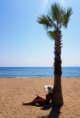Fototapeta na wymiar A woman in white hat sitting under palm tree at the beach