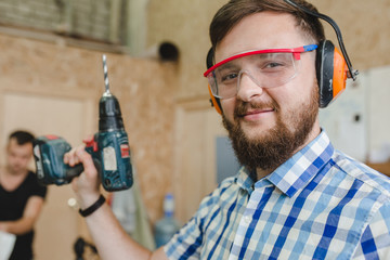 A bearded man in a plaid shirt with anti-noise headphones holds a drill in the carpenter's...